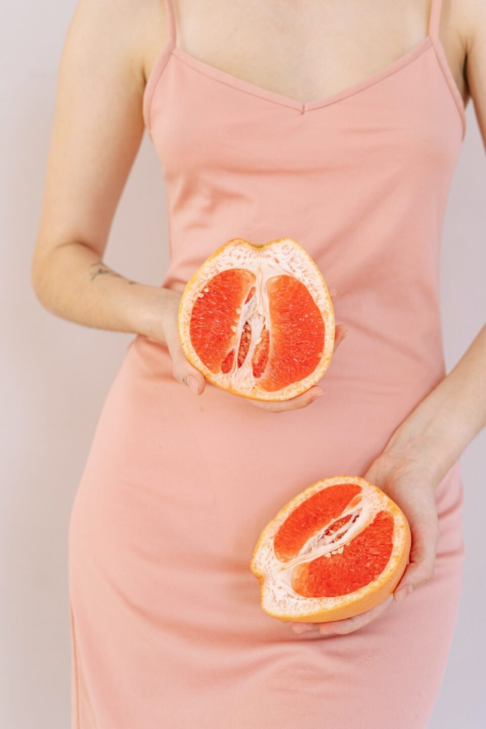 woman in pink sundress holding two halves of cut grapefruit